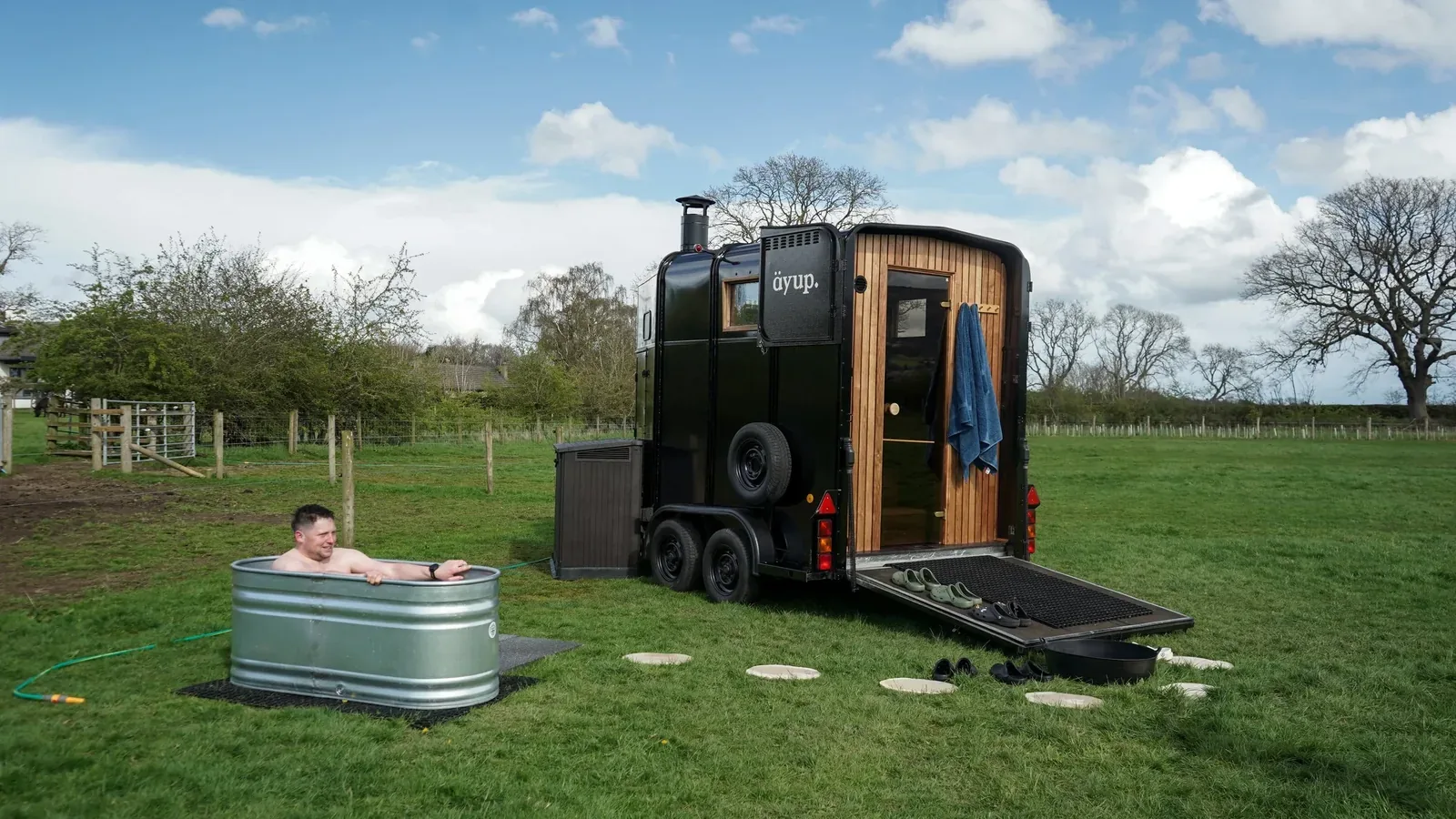 Man sitting in a metal tub of cold water on grass beside a black sauna trailer