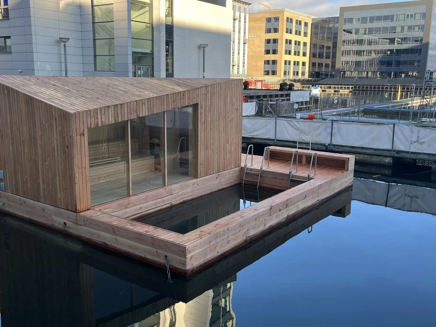 Small wooden floating sauna next to a square swimming area with ladders in a city harbour