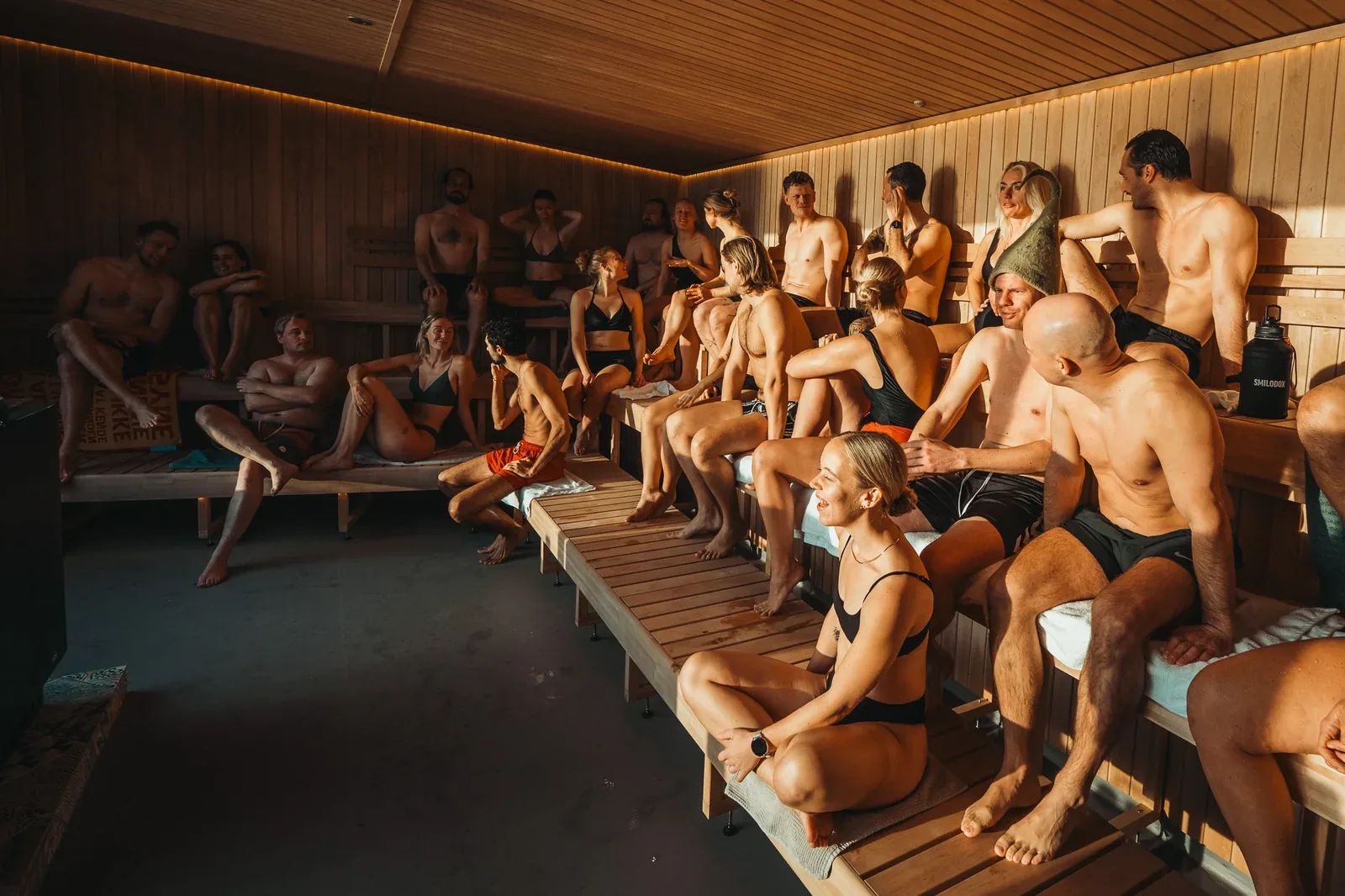 Rows of people in swimwear sitting on wooden benches in a large sauna