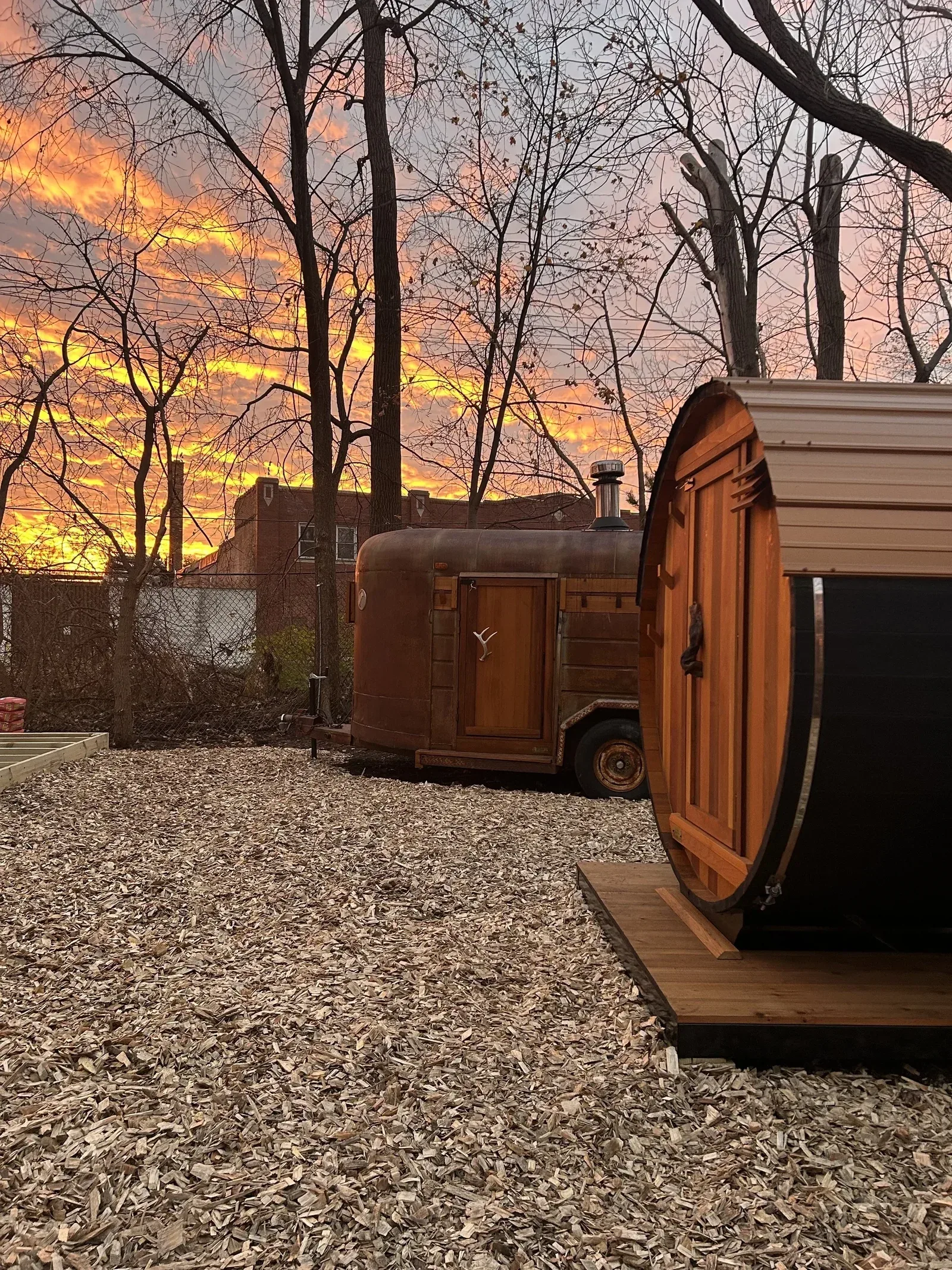 A wooden barrel sauna and a trailer sauna with a chimney at sunset