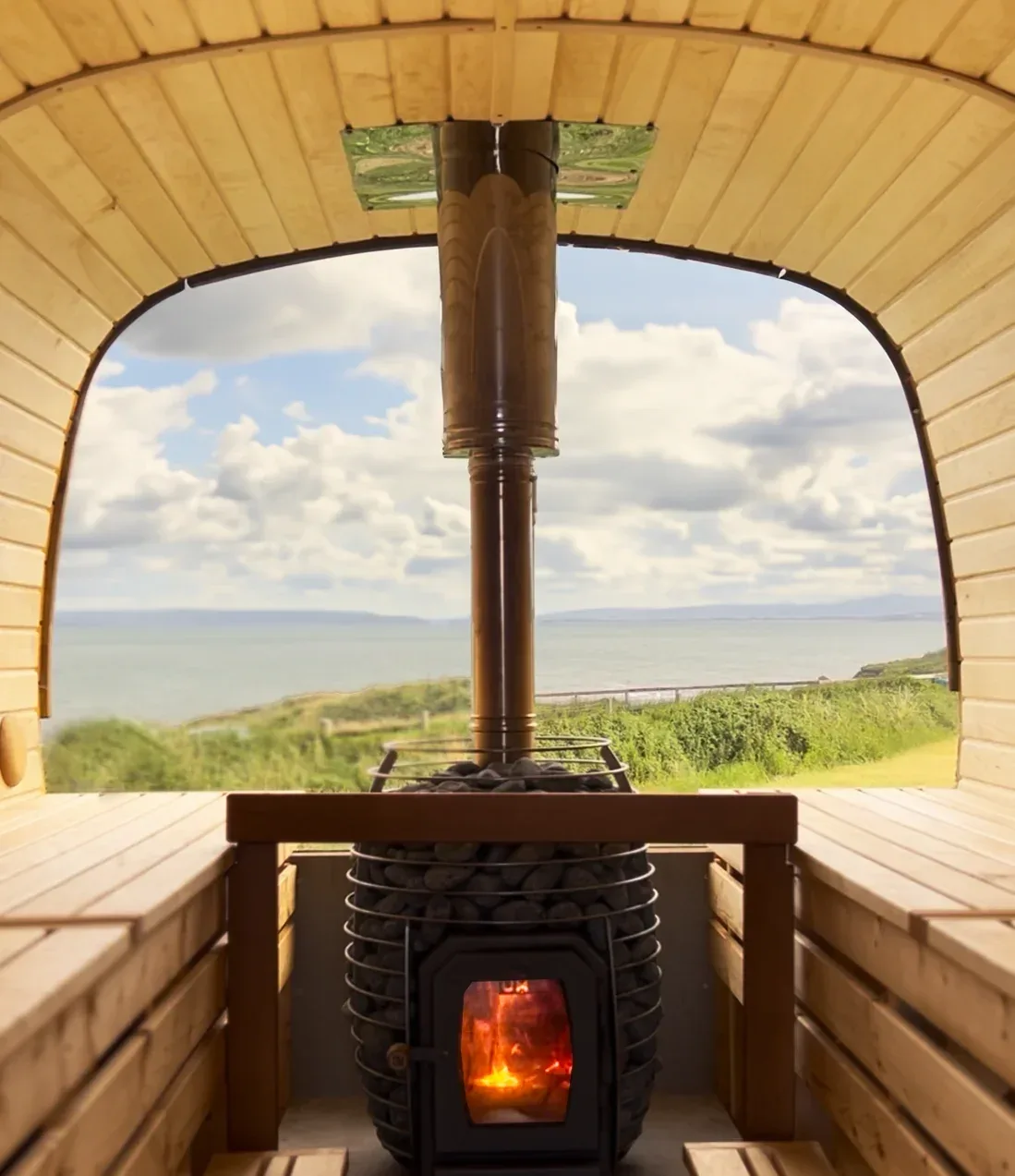 Inside a barrel sauna looking towards a stove and a window with a sea view