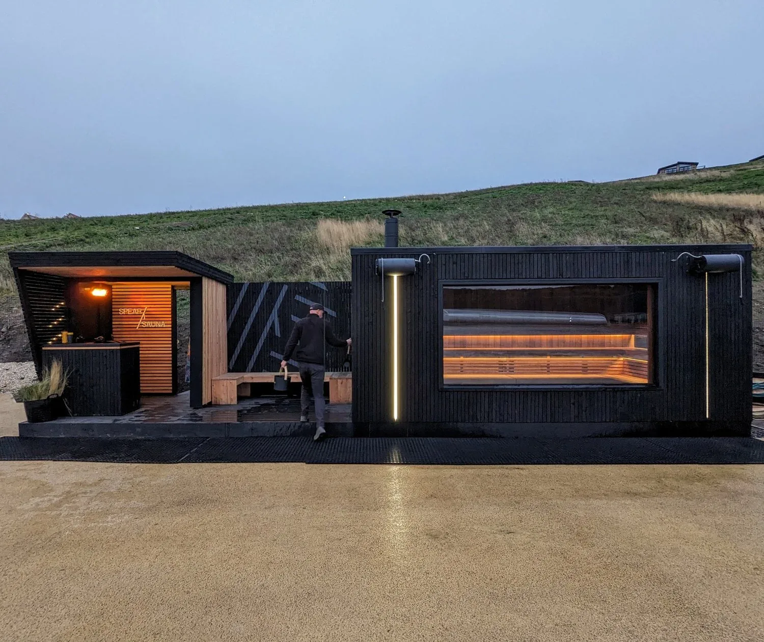 Two black sauna cabins, one with a large window, on an open forecourt