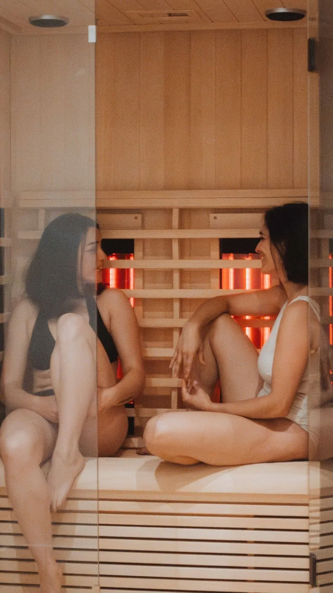 Two women in swimsuits chatting in a small infrared cabin with glowing panels