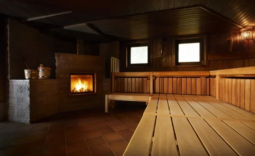 Wooden sauna with long benches and a tiled stove with a fire burning behind glass