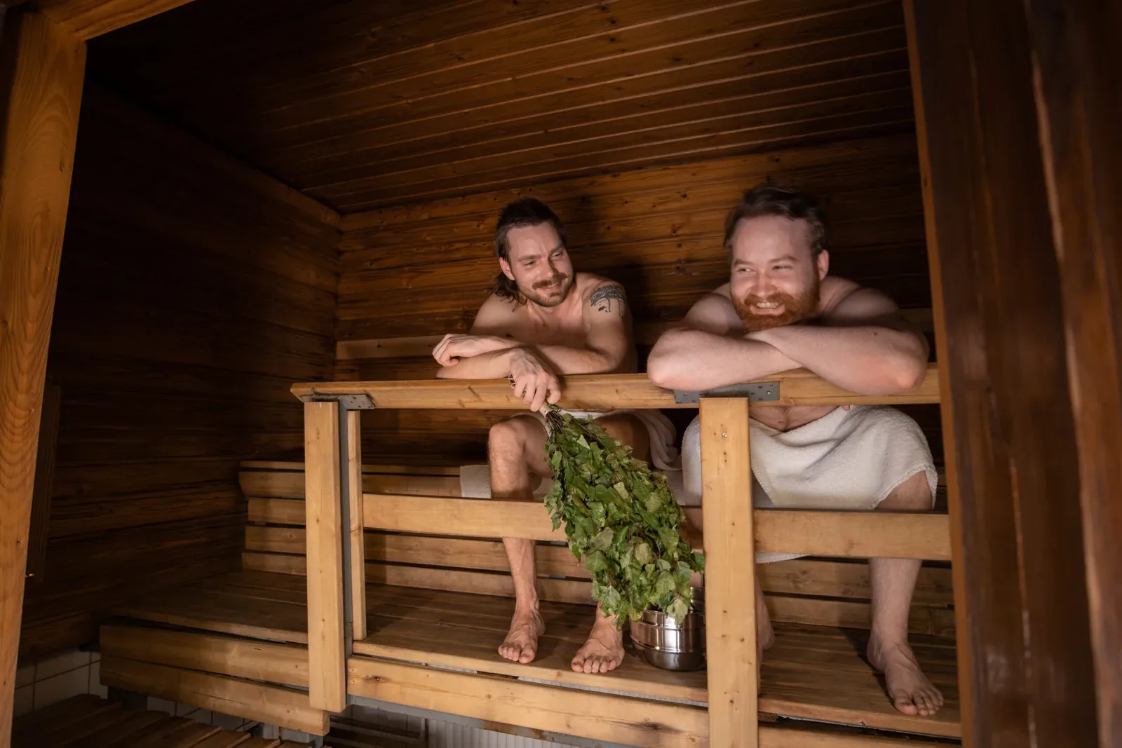 Two smiling men sitting on the top bench of a wooden sauna holding birch whisks