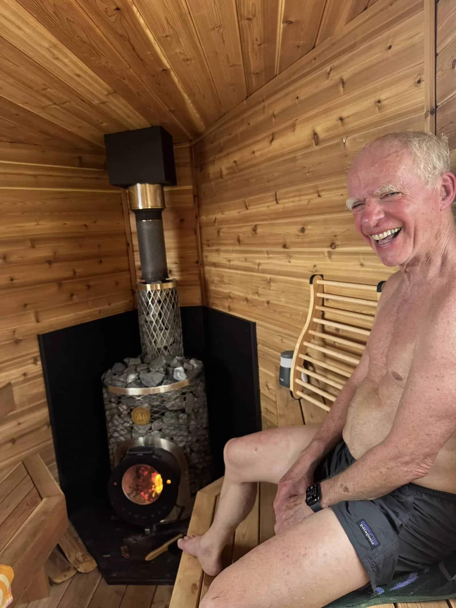 Man smiling on a sauna bench beside a wood-burning stove with a fire inside