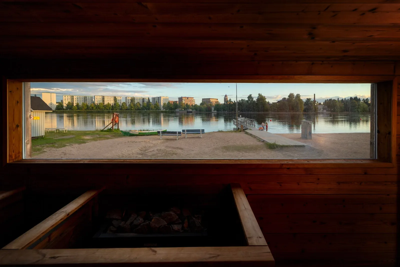 Tmavá sauna s dlouhým úzkým oknem, za kterým je voda, stromy a budovy