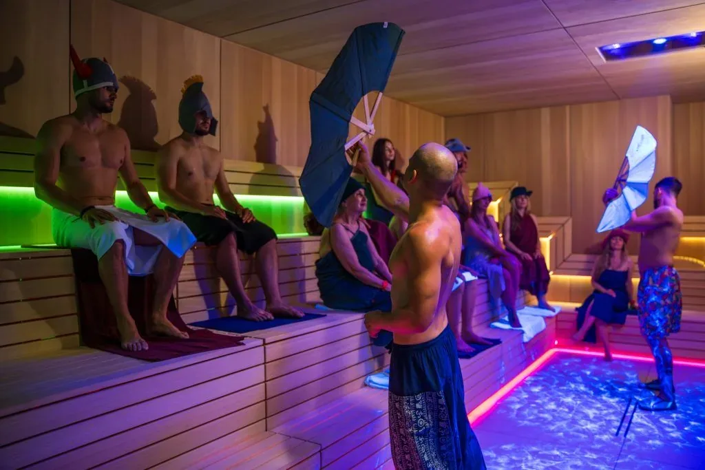 Sauna masters in costume waving large fans in a sauna lit purple and green