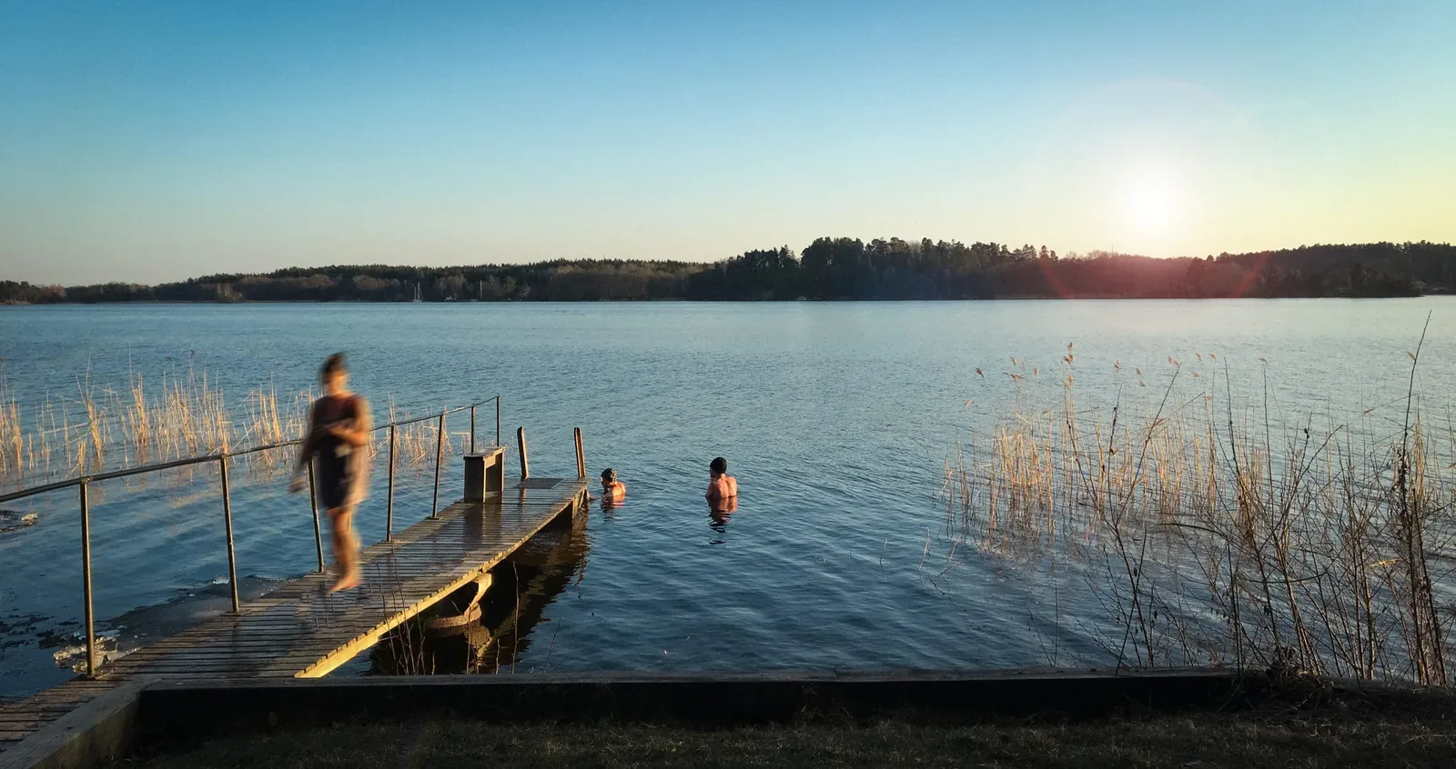 Swimmers in a calm lake beside a wooden jetty at sunset