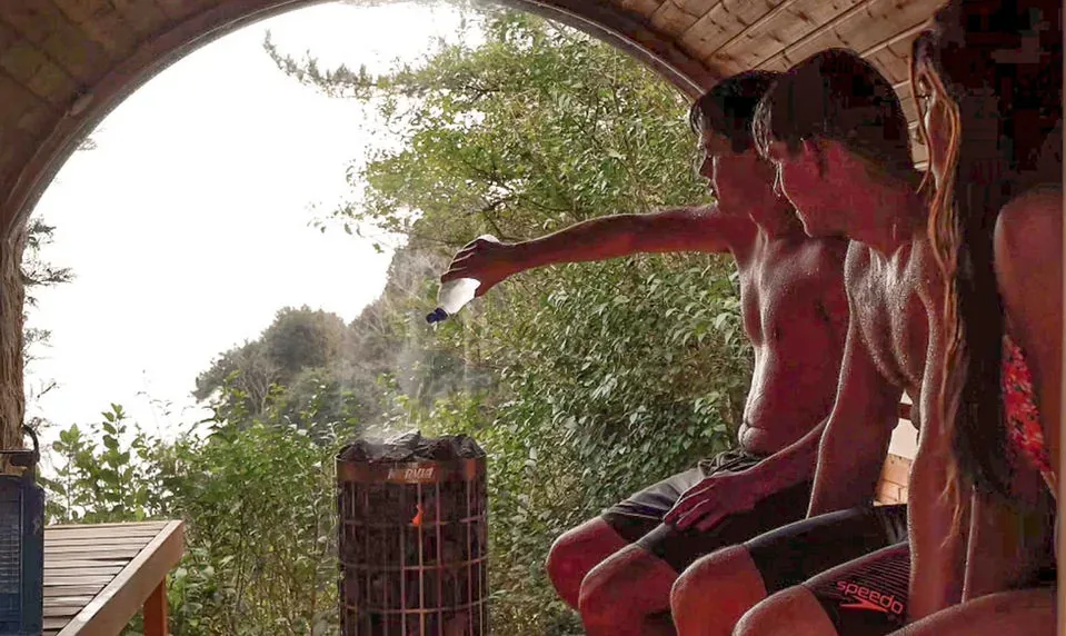 People sitting side by side in an outdoor sauna, one pouring water on the heater