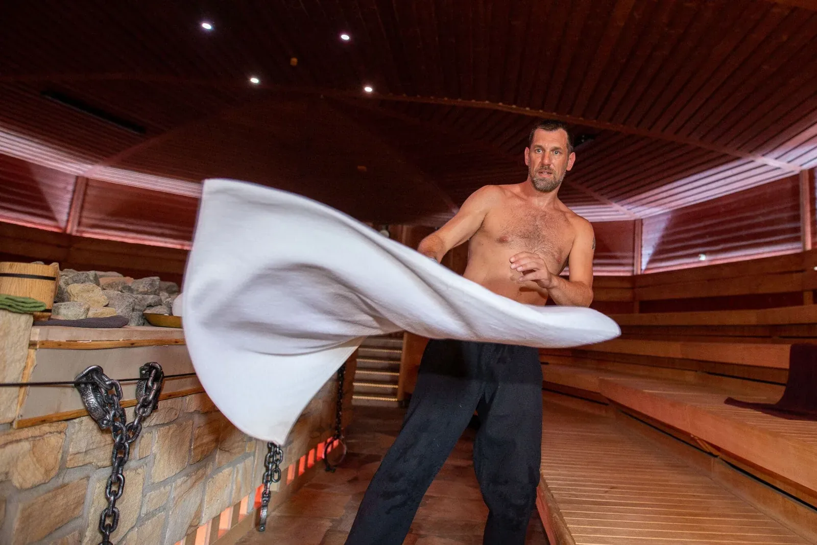 Shirtless sauna master spinning a white towel in a large wooden sauna