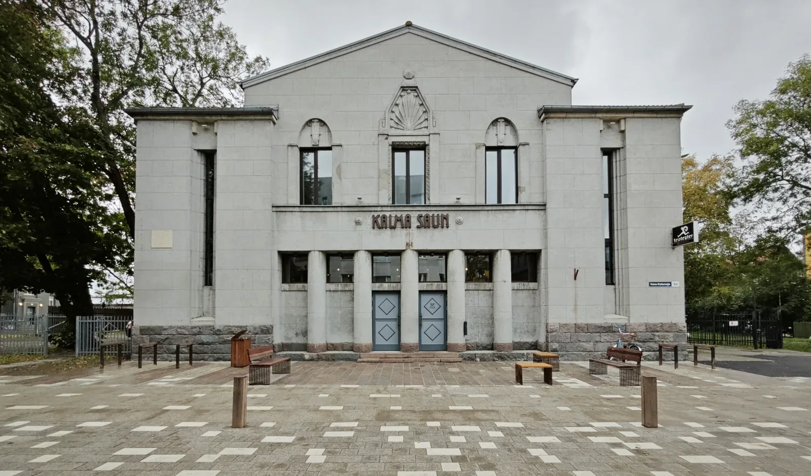 Pale stone building with columns and a sign reading Kalma Saun, with benches in the square in front