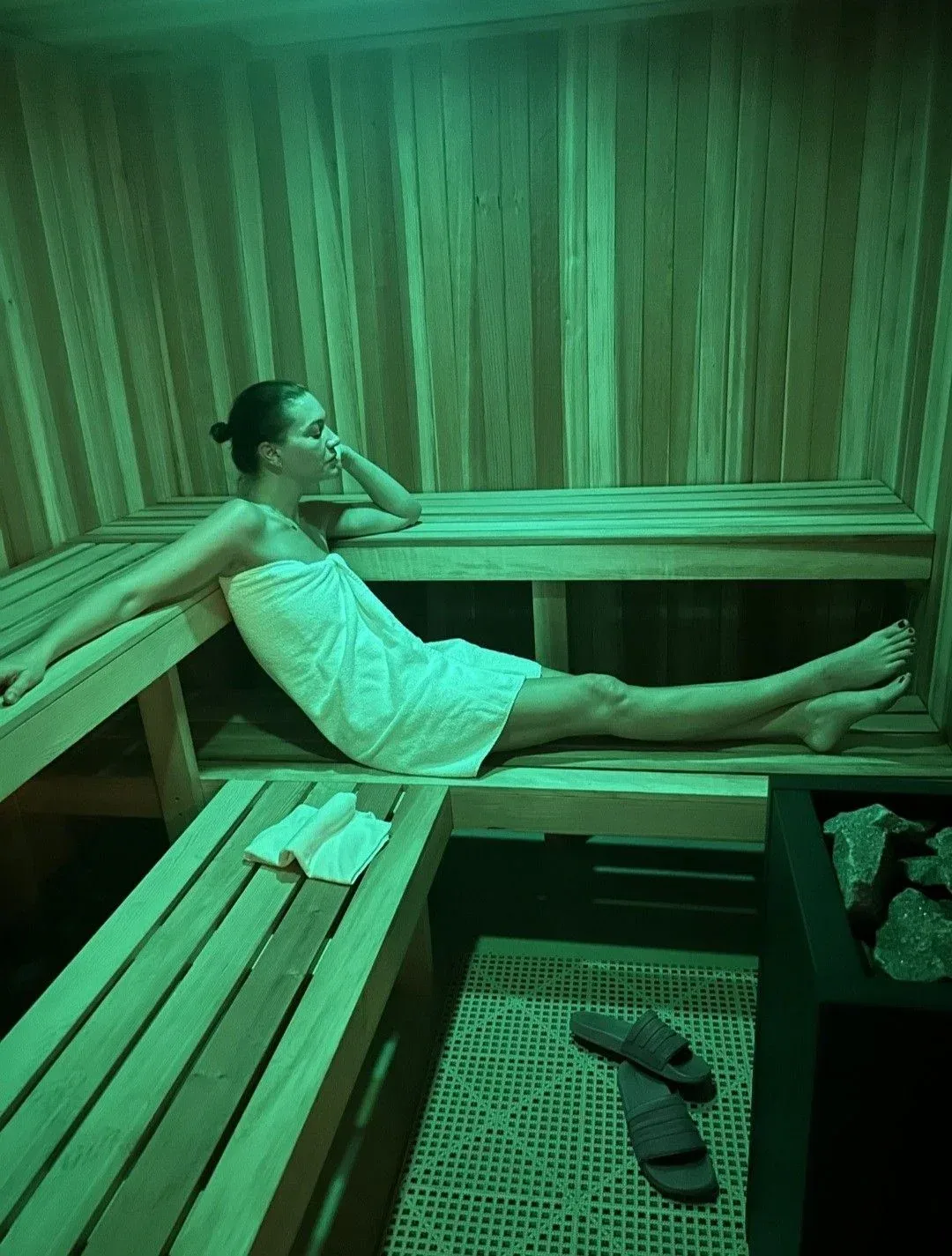 Woman lying on a wooden sauna bench with a towel, flip-flops on the floor below