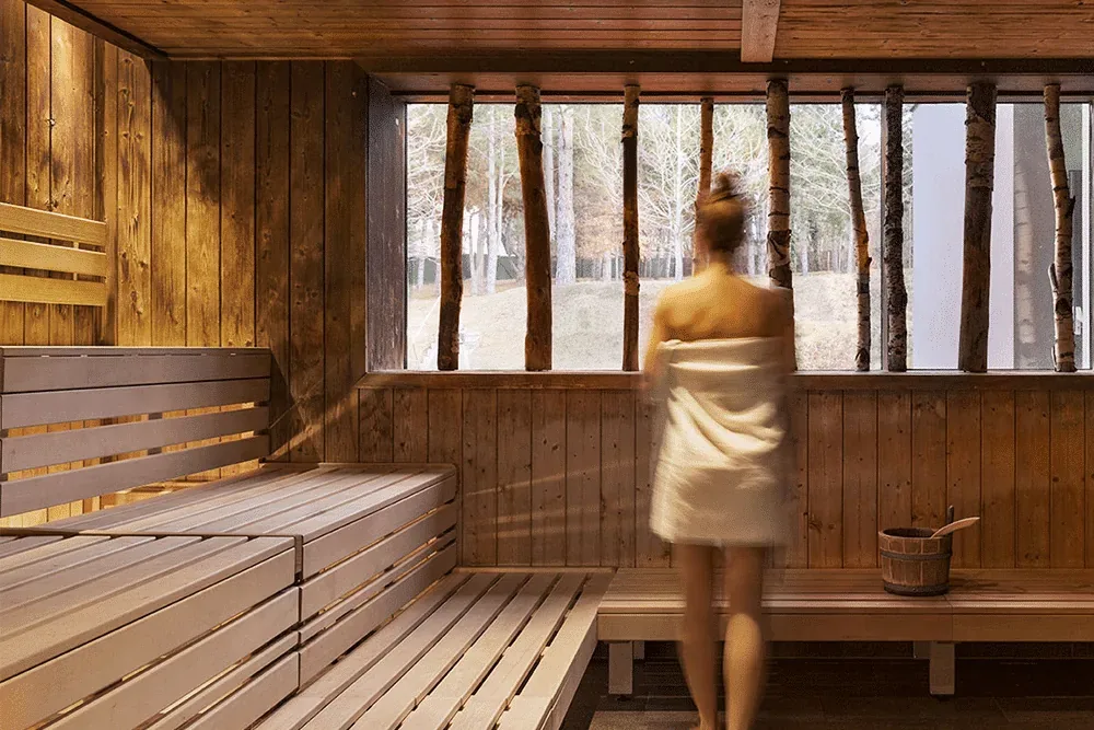 A woman wrapped in a towel standing at the window of a wooden sauna with birch trunks outside