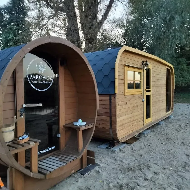 A wooden barrel sauna and a cabin sauna standing on sand under trees