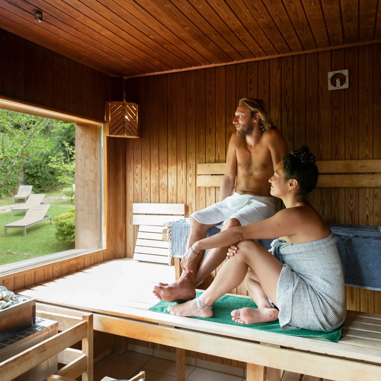 A man and a woman in towels sitting on a sauna bench by a window looking out on a garden